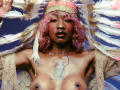 DaniaMarcell with pink curly hair, wearing a feathered headdress, topless with a tattoo below her tits, holding feathers, and a nose ring, against a blue background.