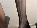 Tattiya's black sheer pantyhose-clad leg with vertical striped pattern, showing subtle light reflection, and a gray shadowed part of a chair on the left side.