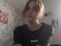 BessBetak has short, messy brown hair and wears a black checkered shirt with a U+Fashion tag, sitting in a dimly lit room with a neutral expression.