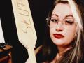 BeccaVanBoa with blonde and dark hair, wearing glasses, red lipstick, holding a wooden guitar with "SIS" written on it, looking slightly to the side.