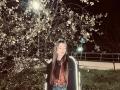 EleanoraMirabal with long brown hair, eyes closed, wearing a black jacket, red plaid shirt, and blue jeans, standing in front of a blooming tree at night.