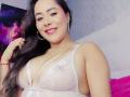 CamilaBardot with long black hair, smiling, wearing white lace lingerie that reveals her big tits, pink lipstick, and silver earrings, in a bright room.