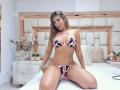 KendraMiler with tan skin, long brown hair, wearing a cow print bikini with pink straps, sitting on a bed, knees apart, showing her tits and pussy.