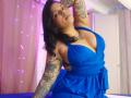 Chili with tattoos, blue dress, large boobs, arm raised, black wavy hair, confident expression, pink and purple lights in background, standing in bedroom, looking at camera.