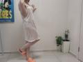JoannaSia in a sheer white dress, standing on one leg with her ass slightly raised, wearing orange slippers, in a room with a potted plant and a poster.