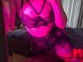 Milagres is wearing black lace lingerie with straps, showing her tits and ass. She's kneeling in pink-tinted light, reflecting in a mirror, with a bed in the background.