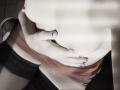 Les seins d'OrianaLaFrancaise, couverts par un tissu rouge, sont doucement maintenus par sa main dont les ongles sont peints. Elle porte des bas noirs en dentelle qui montent jusqu'à la cuisse. Une lumière douce et ombragée..