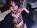 ViolettClay with dark hair, black lipstick, and eye makeup, wearing a black mesh lingerie set with cutouts, black choker, and open robe, sitting on a blue bed.