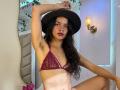 LucyySky with long black hair, wearing a maroon lace bra, black hat, and no panties, showing armpit hair, sitting with arm raised, and red lipstick, looking at camera.