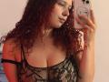 ValeryScot with curly red hair, wearing a black lace top, showing her tits, taking a mirror selfie with a phone. She has a necklace and a tattoo on her arm.