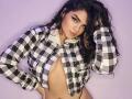 CandyLatina69 with curly dark hair, wearing an unbuttoned black and white checkered shirt, exposing her tits, against a purple background, looking seductively at the camera.
