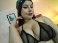 Melani Becker con seni grandi e evidenti, indossa un reggiseno nero trasparente, ha l'ombretto rosa, una bandana e rossetto rosa, una mano che tocca il viso, sorride leggermente..