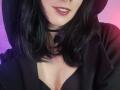 IssisDiosa has black shoulder-length hair, wearing a black hoodie with a low neckline, showing a bit of her tits, and a black choker with a small pendant.