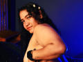 JoyceHill, topless, with curly black hair, small white flowers, and a nose ring, smiling, showing a tattoo on her right arm, and wearing a black wristband, in blue light.