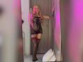 MiaValenttine with long blonde hair, lace black lingerie, red garter, tattoos on arm and thigh, taking a mirror selfie, pink and purple lighting, standing in a room.
