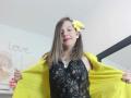 ArianaBlonde with medium tits, wearing a black lace top and yellow flower in her brown hair, smiling, holding a yellow jacket open, red lipstick, in a well-lit room.
