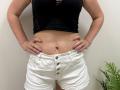 Tilliana with hands on white denim shorts, black lace-up top, pink nails, showing her flat stomach and navel against a plain wall, with a green plant on the right.