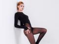 GrettaDemichelis, with red hair, wearing a black long-sleeve bodysuit and sheer black tights, leans against a white wall, hand on her butt, legs apart, looking at camera.