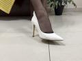 Rorria's feet in sheer black pantyhose with white pointed-toe stilettos, standing on a shiny white tile floor, heel visible, yellow fabric in the background.