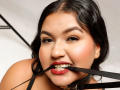 DojaAissha has black hair, red lipstick, and a black strap in her mouth with a collar around her neck. Her eyes are open, looking at the camera.