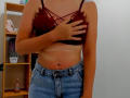 Steisii with tan lines, wearing a sheer red bra, covering her small tits with one hand, and blue high-waisted jeans, standing in a room with a wooden shelf.