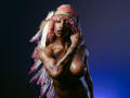 DaniaMarcell with pink and white feathered headband, topless, showing her big boobs and tattoos, dark skin, hand near face, blue and dark background. Her nipples are visible.