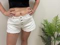 Tilliana's bare belly with a navel, wearing white frayed shorts with buttons, black top, pink glitter nails, hands on hips, standing next to a green potted plant.