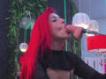 AlissaBrown with bright red hair, wearing a black top, sucks a dildo into her mouth. She's holding a microphone, looking up, with green and white lights in background.