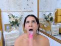 AmaiaCooper's face covered in cum, mouth open, tongue out, with a pink dildo in front. Dark hair, brown eyes, earrings, white wall with gold decor, yellow chair.