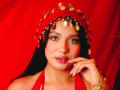 LilyConorr with shiny green eyes, glossy pink lips, and dark hair, wearing a red headpiece with gold coins, touching her lip with a gold bracelet on her wrist.