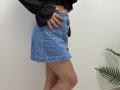 Tilliana's blue denim skirt with buttons, black long-sleeve top, pink nails, and tan skin, standing with her hand on her hip, showing her bare legs.