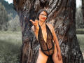 ParisMaturre with glasses, wearing a black lace bodysuit, standing against a tree, pointing with her right hand, her tits visible through the lace, in a grassy field.