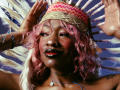 DaniaMarcell with pink curly hair, nose ring, and tribal headband, hands raised, feathers in background, shiny black lips, red lipstick, tattoos, and blue bracelet, looking confident.