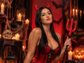 AriiaMon with long black hair, red devil horns, red corset, big tits, holding a red trident, surrounded by candles, skull, and Halloween decor, looking sexy and confident.