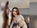 MandyMouses con lunghi capelli castani, che indossa un top corto bianco trasparente e biancheria intima floreale, inginocchiata su un letto, che mostra i seni e il sedere, in una camera da letto..