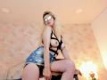 SophieTeller with blonde hair, wearing black strappy top exposing tits, blue denim skirt, black panties, reflective sunglasses, sitting on table, light skin, room with pink textured wall.