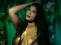 MariaRowe with long black hair, wearing a glittery gold top, exposing large breasts. She's looking seductively at the camera, hand in her hair, red lipstick, green eye shadow. Dark, moody background.