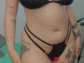 AmberLaurent with tan skin, black bra, and strappy black panties, showing tattoos on arms and thighs, a belly button piercing, and pink butt plug visible.
