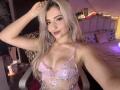 EliseMonet, avec de longs cheveux blonds, un soutien-gorge en dentelle rose laissant entrevoir ses seins, du rouge à lèvres rose, une main dans les cheveux, souriante, un lit rose, des coussins à motifs, un éclairage tamisé, assise sur une chaise..