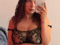 ValeryScot with curly brown hair, wearing a black lace bra, showing her tits, holding a phone, red tattoo on right hand, fair skin, pink lips, in a mirror selfie.