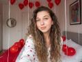 Sanella with curly brown hair, wearing white heart-print shirt, red balloons hanging, smiling, on bed with red blanket, white pillows, and colorful wall art.