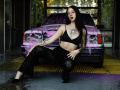 AlexLovely has black hair, wearing a black bra and lace pants, sitting in front of a purple-sprayed car with tattoos, including a sun on her chest, and dark lipstick.