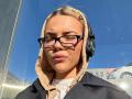 ArnettaNazario with closed eyes, black glasses, beige hood, black headphones, and a black jacket, sunlight on her face, blonde hair, blue sky reflection.