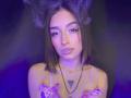 LunaixMoon with smoky hair, pink bowed top, lip ring, necklace, and tattoo, staring seductively, purple-blue lighting. Her tits are small, and she has a cute face.