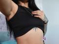 LuciadelaVega's small tits are covered by a black ribbed crop top, revealing a tattoo below her right boob. She has long black hair, a gold necklace, and tattooed arms.