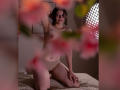 MaryCruzz kneeling on a bed, topless with small tits, wearing beige panties, short curly brown hair, tattoo on right shoulder, soft light, blurred pink flowers in foreground.