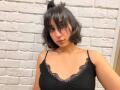HeikeCarchi with short black hair, cat ears, wearing a black lace-trimmed tank top, showing her small tits against a white brick wall. She has a neutral expression.