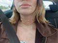 MissFrenchy's face in a car, wearing a brown jacket, gold necklaces, and a seatbelt, with light brown hair and glossy pink lips, visible from the chin down.