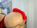 Les fesses de NaomyHott69 en lingerie rouge avec une queue fourrée rouge, penchée en avant, montrant un tatouage en dentelle sur le bas du dos, des cheveux noirs bouclés et un oreiller jaune..