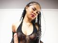 LuciaVargasX with braided hair, wearing a sheer black mesh top, exposing her small tits, holding a big, uncircumcised, erect cock, wearing gold earrings, red lipstick, and a necklace.